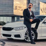 bearded male in sunglasses dressed in a black suit sitting on luxury car against a skyscraper.